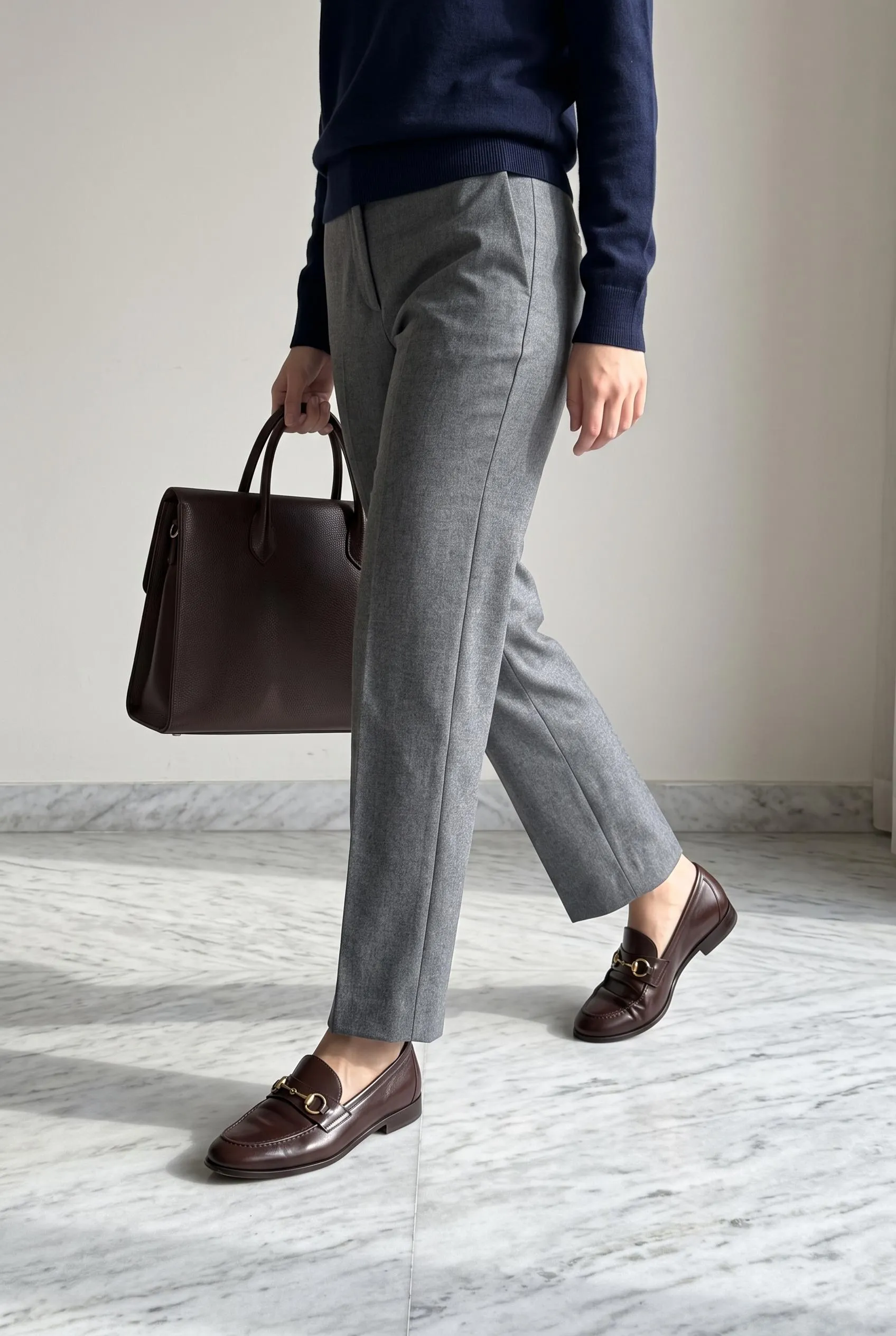 Brown horsebit loafers with grey wool trousers, a navy knit and a brown work bag