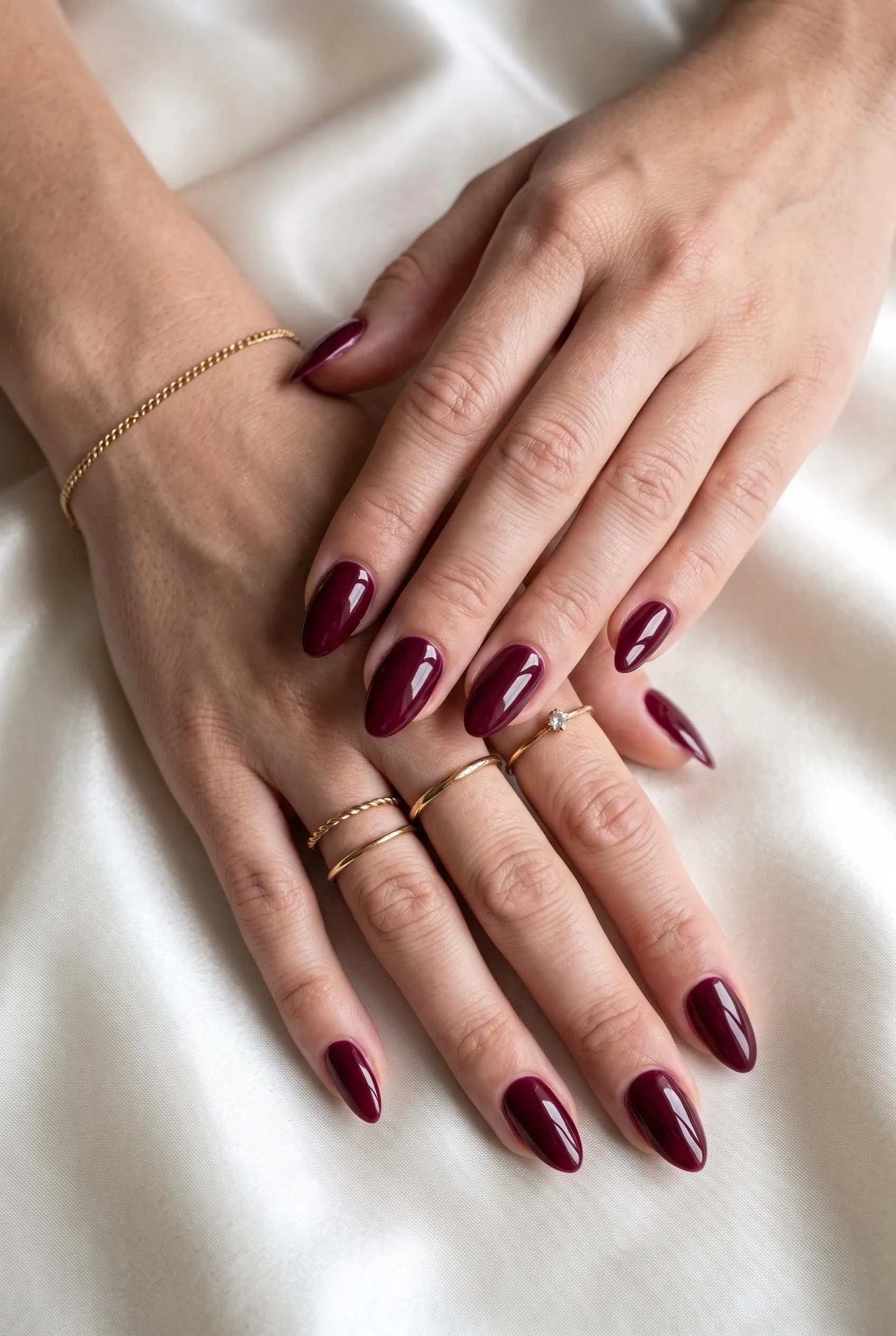 Glossy deep burgundy almond nails on two crossed hands on ivory satin