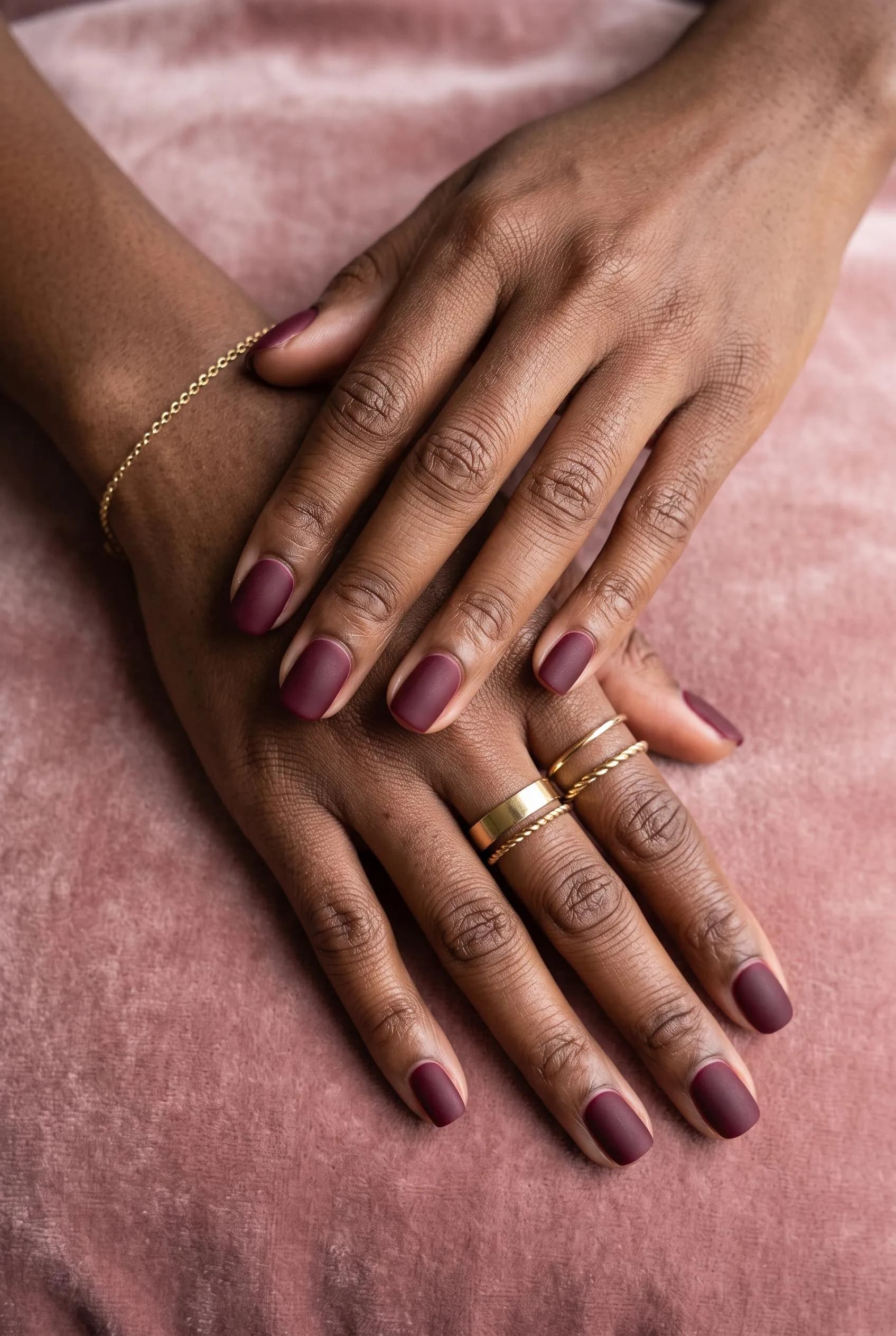 Short squoval matte burgundy nails on deep brown hands on dusty-rose velvet