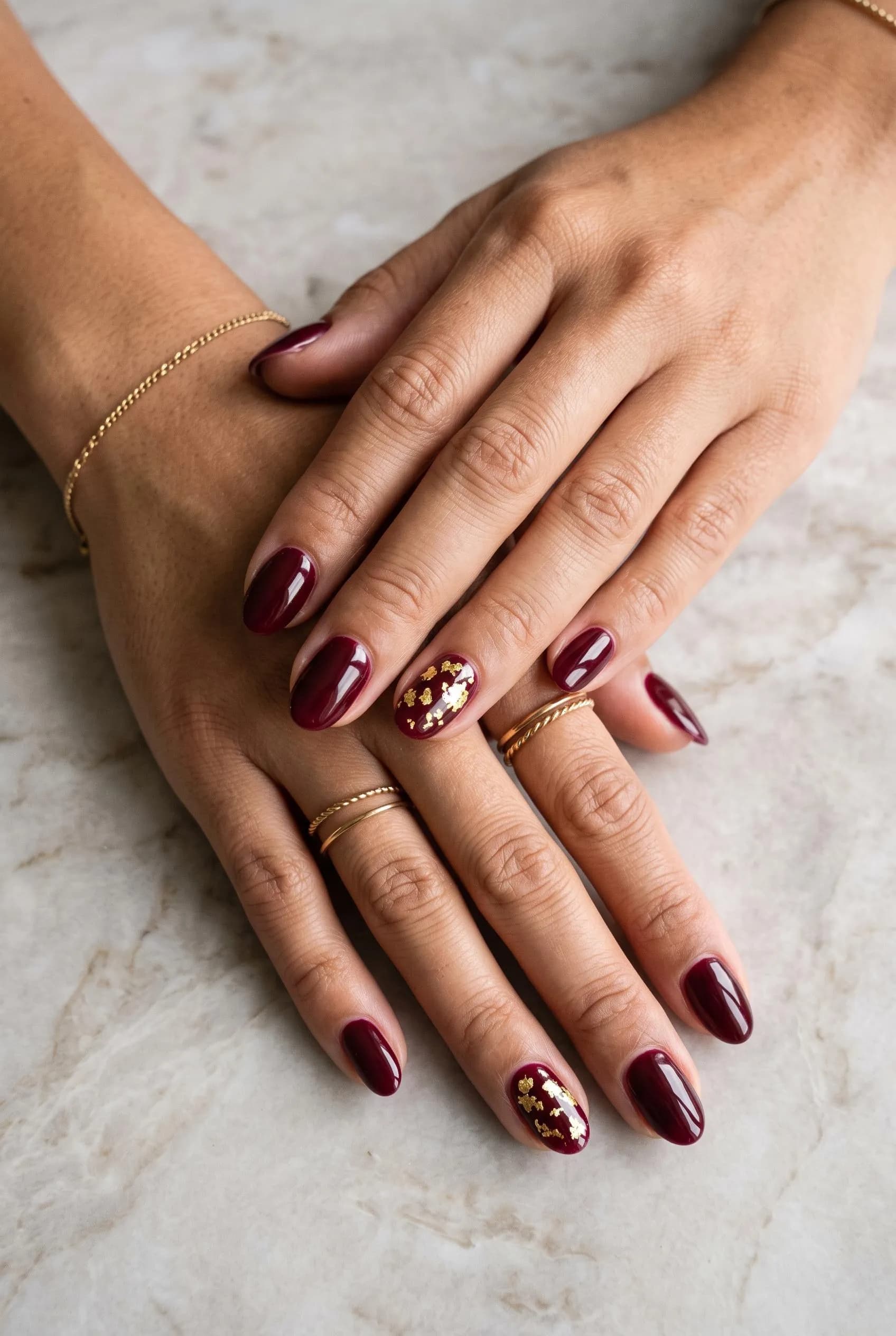 Glossy burgundy nails with gold leaf foil on the ring fingers on marble