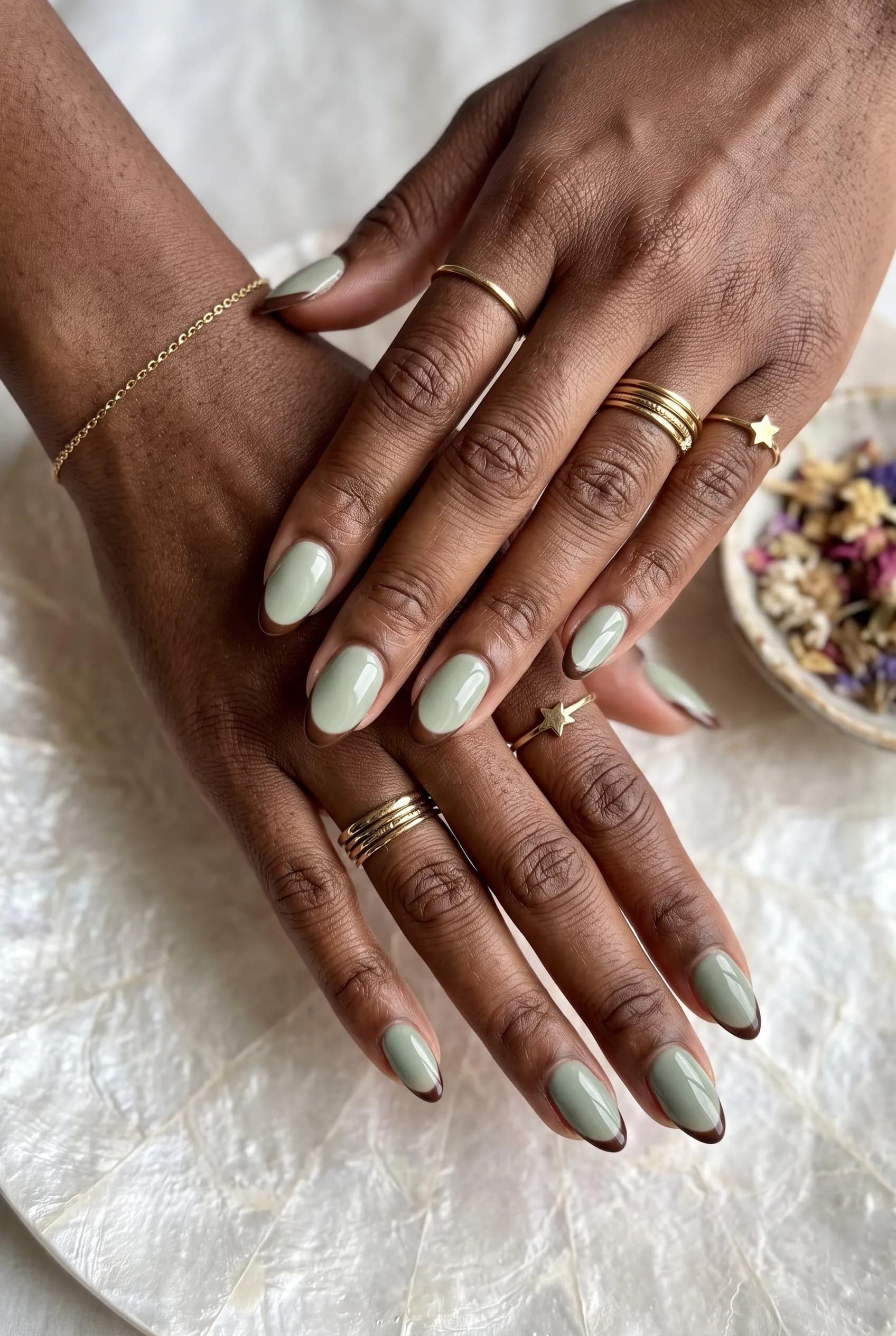 Sage green almond nails with thin chocolate brown French tips on deep brown skin