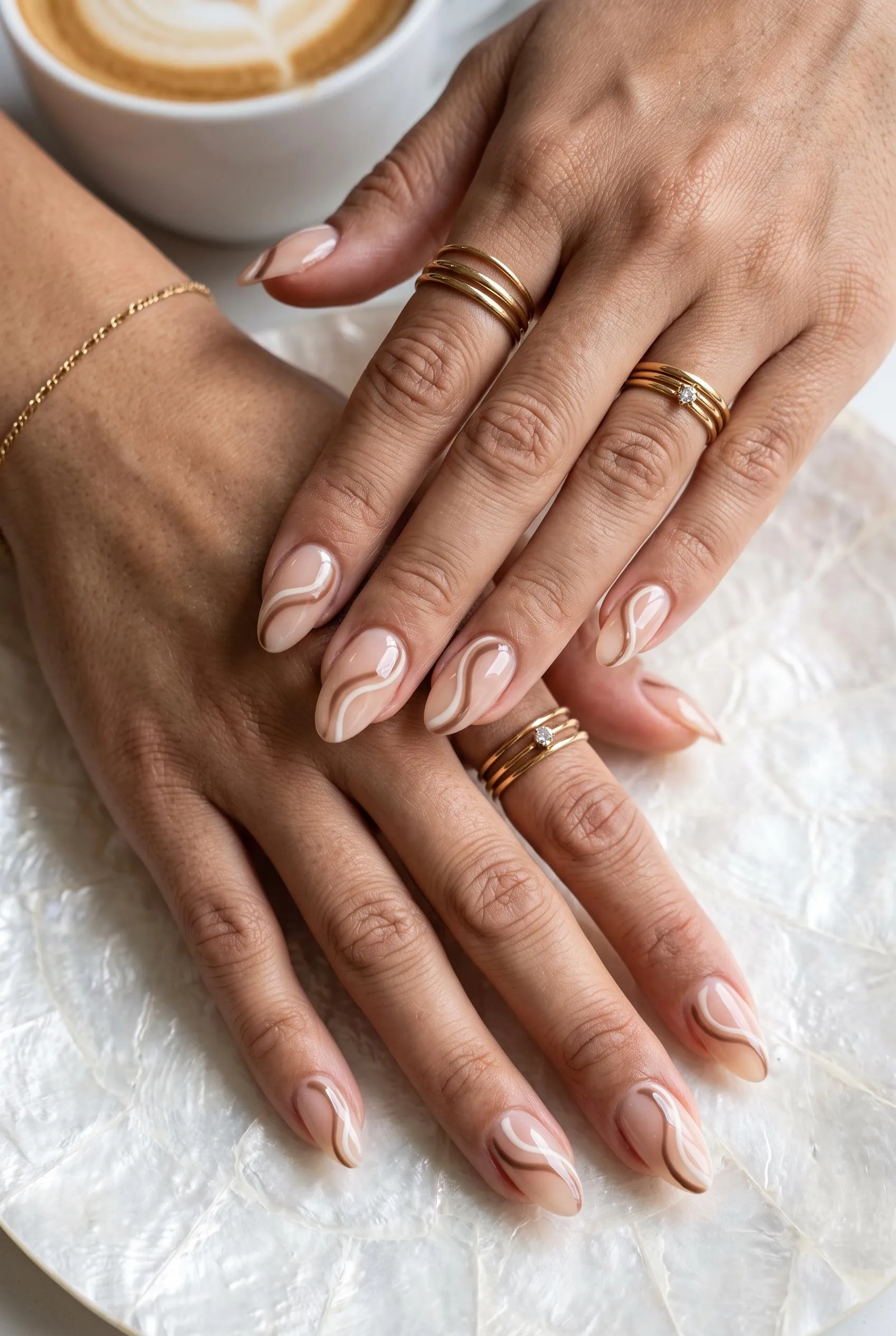 Sheer nude nails with thin mocha and cream swirls beside a cappuccino