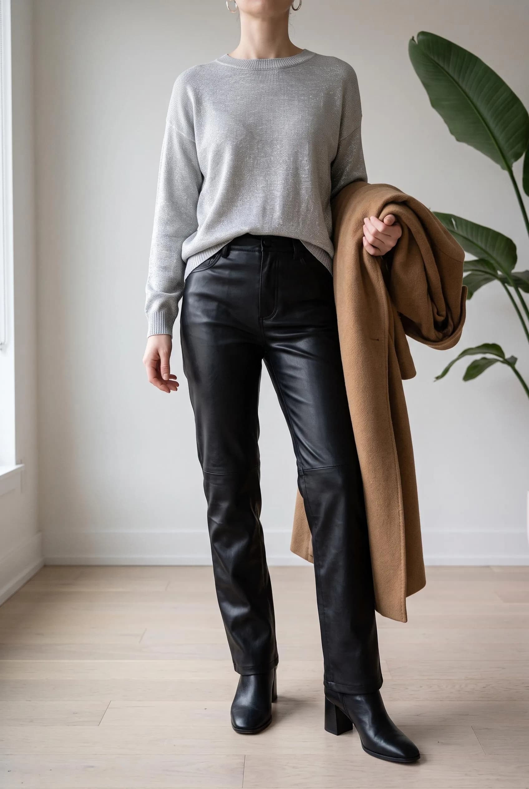 Grey knit sweater with black leather trousers, ankle boots and a camel coat over one arm