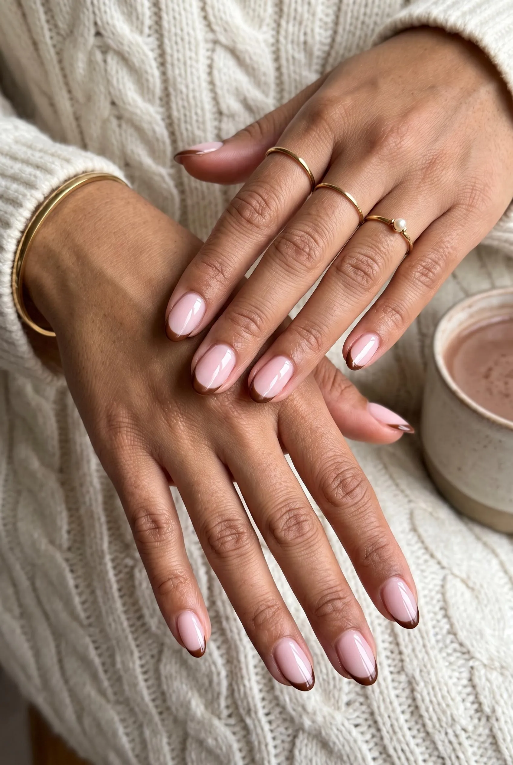 Sheer milky pink oval nails with thin chocolate brown French tips on a cream cable knit