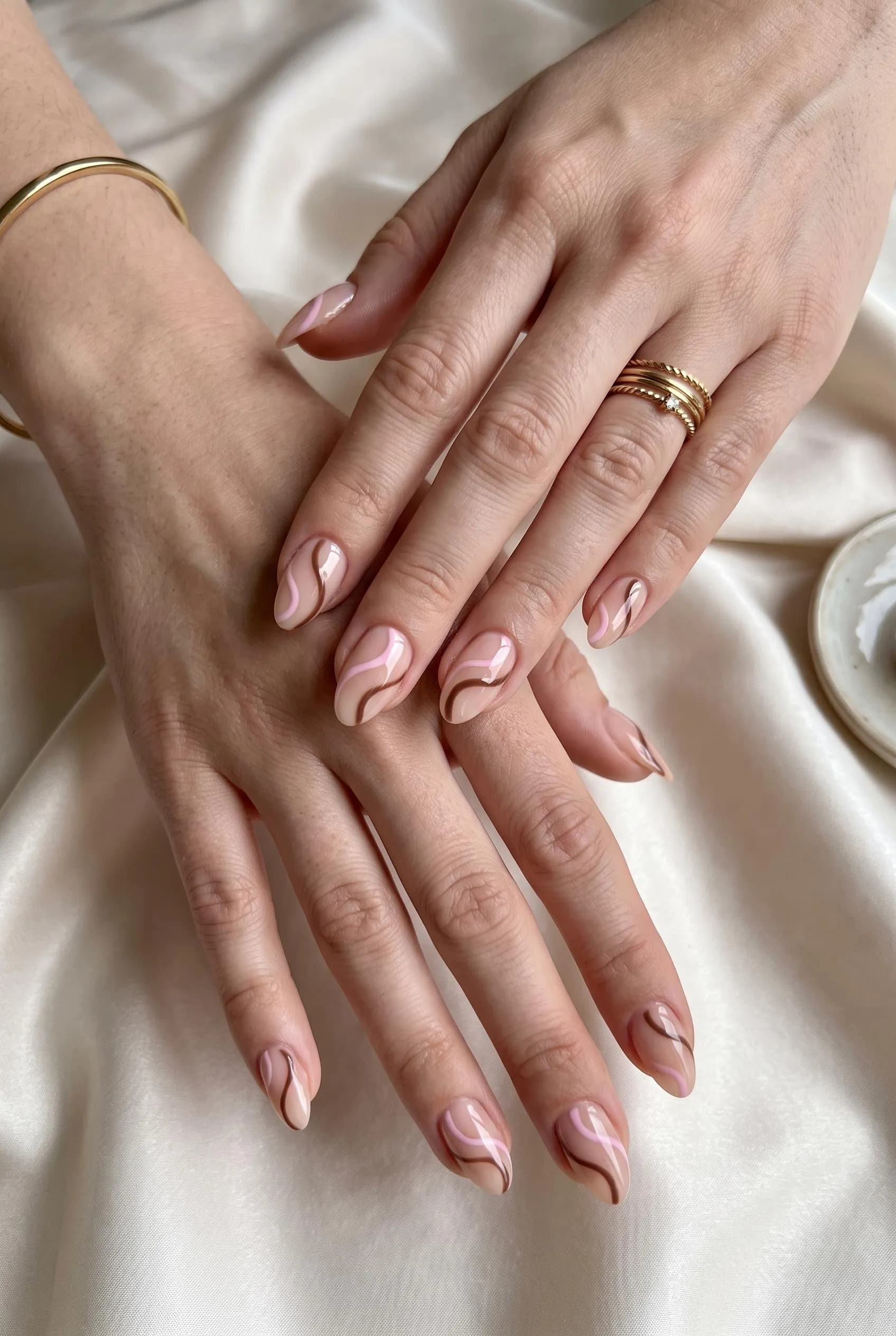 Sheer nude almond nails with thin pink and brown abstract swirls on ivory silk