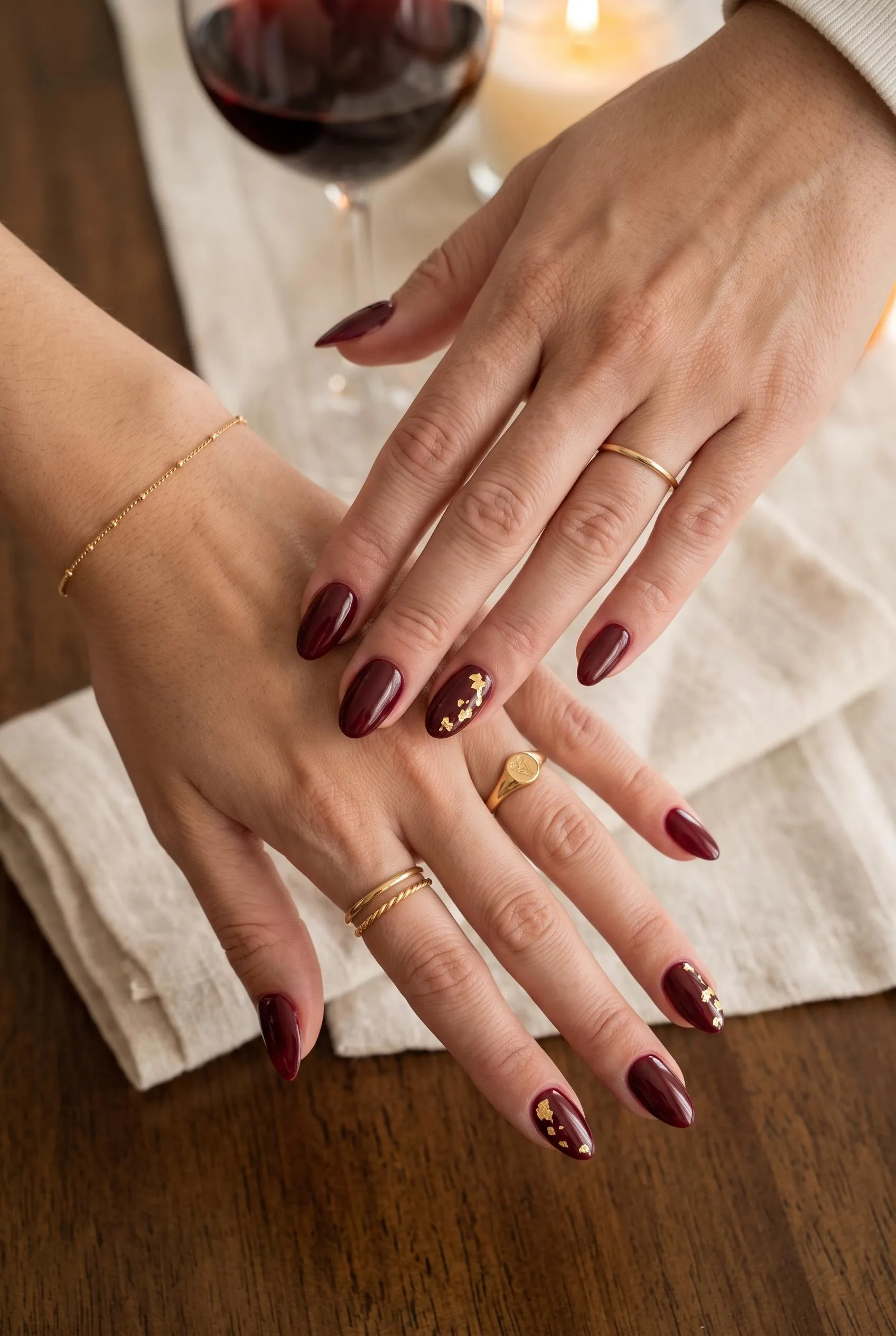 Glossy burgundy almond nails with gold foil on the ring fingers beside a glass of red wine
